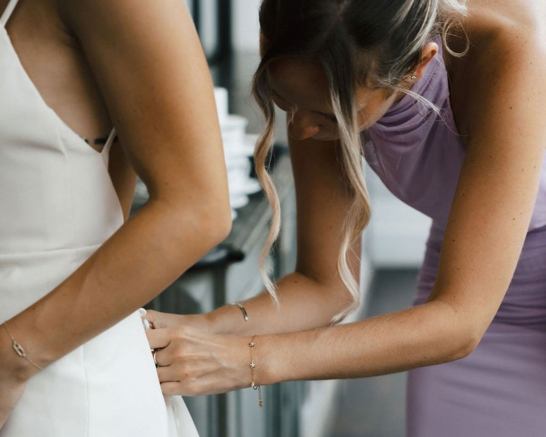 A bride in a white wedding gown has a bridesmaid in a lavender dress adjusting the skirt. The bride is wearing delicate gold chain bracelets, and the bridesmaid is wearing a gold chain bracelet with a cross pendant.