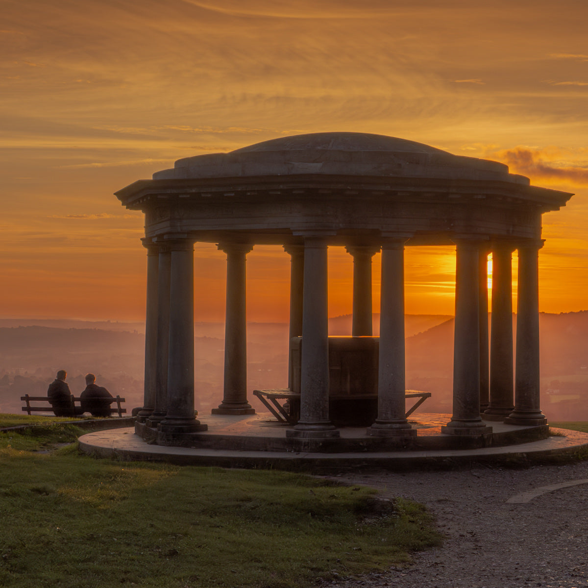 A classical stone pavilion with tall columns stands on a grassy hillside at sunset, with two people sitting on a bench to the left. The sky is filled with warm orange and yellow clouds, and the sun is setting behind the structure.