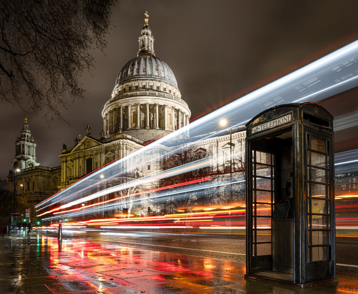 London night scene with St. Paul's Cathedral dome in the background, long-exposure light trails from passing cars across the street, and a classic black telephone booth in the foreground.