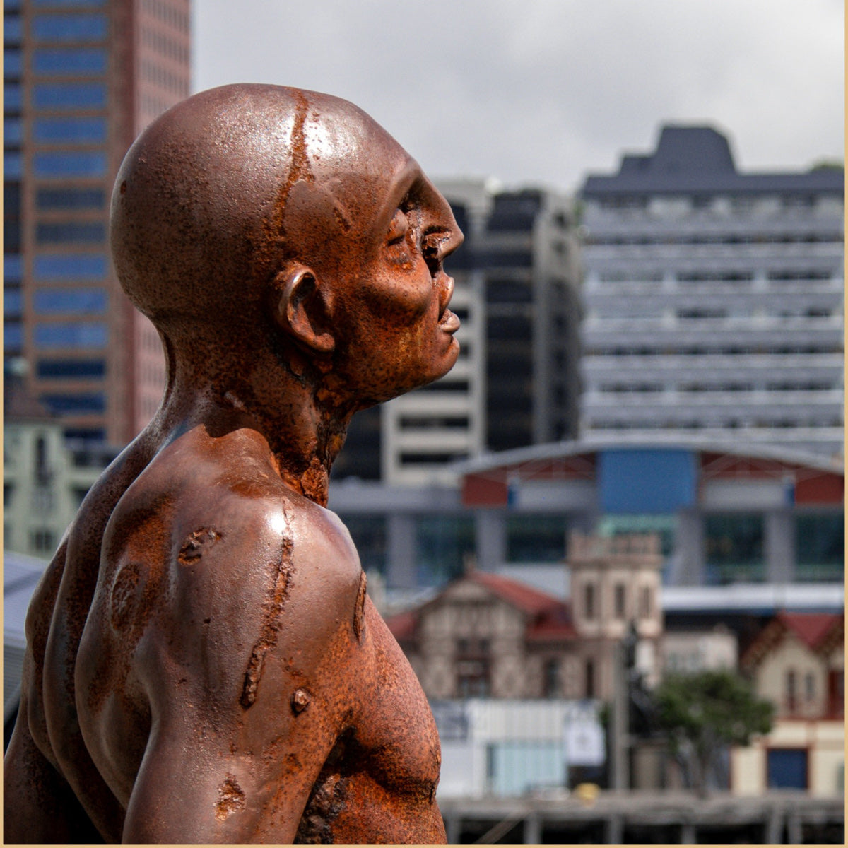 A close-up of a rusted metal sculpture of a bald man's head and upper torso in profile, set against a blurred city skyline background.