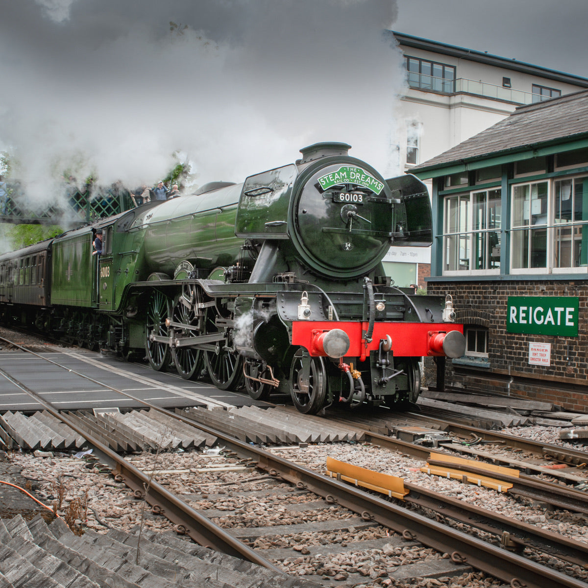 A green steam locomotive train with a red front buffer is positioned on railway tracks beside a station building labeled REIGATE. Steam billows into a cloudy sky, and the station architecture and tracks fill the scene.